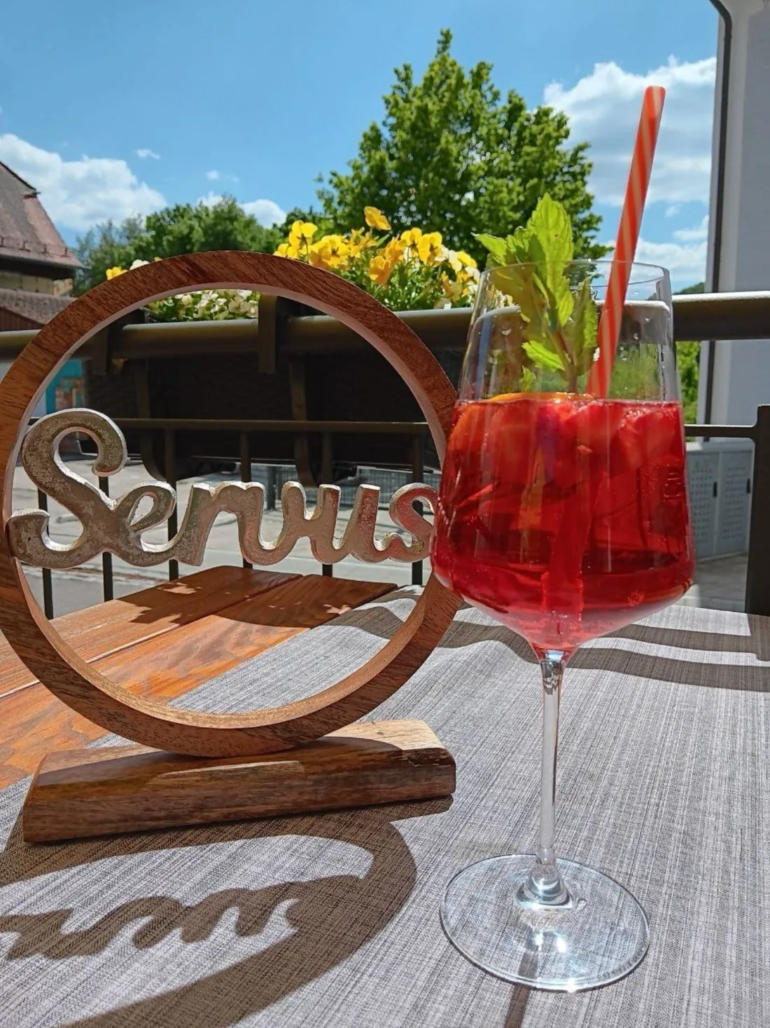 Geschnitztes Servus-Schild aus Holz neben einem roten Spritz auf der Terrasse